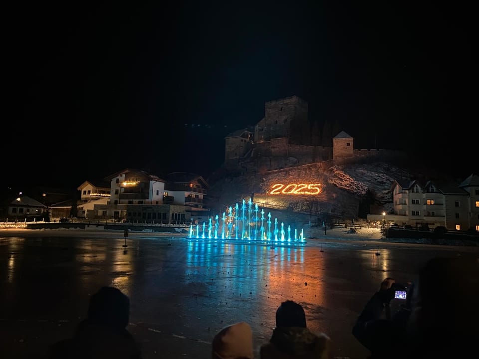 Nächtliche Winteraufnahme eines beleuchteten Brunnens vor einer Burg auf einem Hügel.